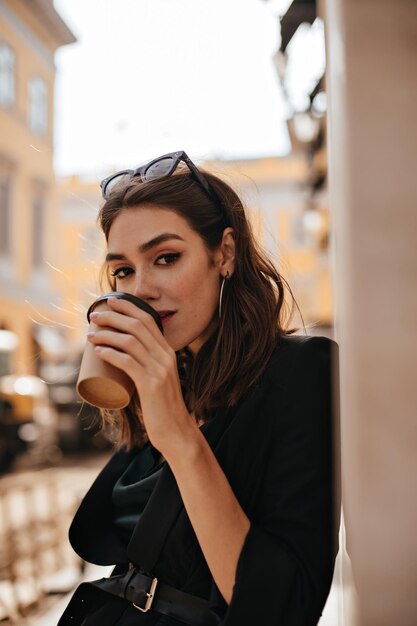 Elegante joven con cabello ondulado morena maquillaje moderno y chaqueta negra bebiendo una taza de café en la terraza del café de la ciudad y mirando directamente