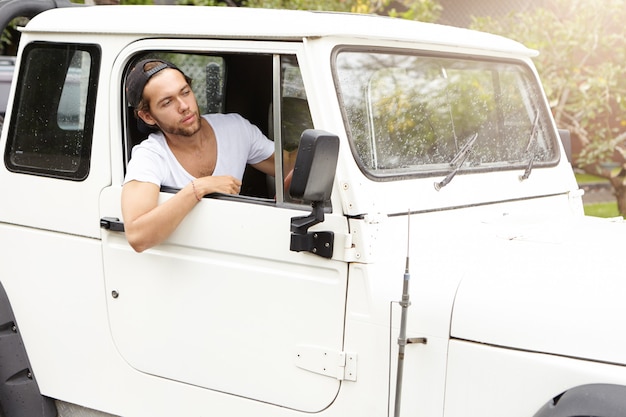 Elegante joven caucásico mirando por la ventana abierta de su vehículo utilitario deportivo blanco. Hombre sin afeitar con gorra de béisbol al revés conduciendo su jeep, disfrutando de un viaje por carretera