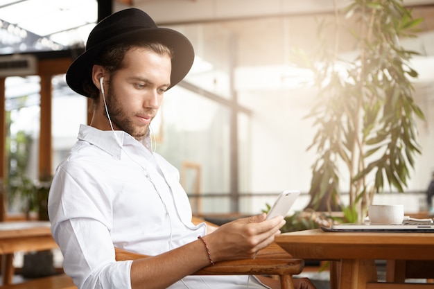 Elegante joven caucásico con sombrero negro enviando mensajes de texto sms o leyendo publicaciones a través de las redes sociales usando wifi gratuito en su teléfono móvil durante el desayuno en la acogedora cafetería y escuchando música en auriculares