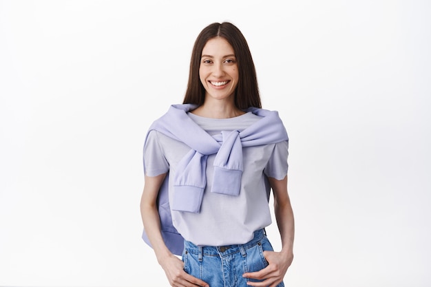 Elegante joven delgada en traje de estilo de vida, tomados de la mano en los bolsillos y sonriendo al frente, de pie informal en pose relajada contra la pared blanca