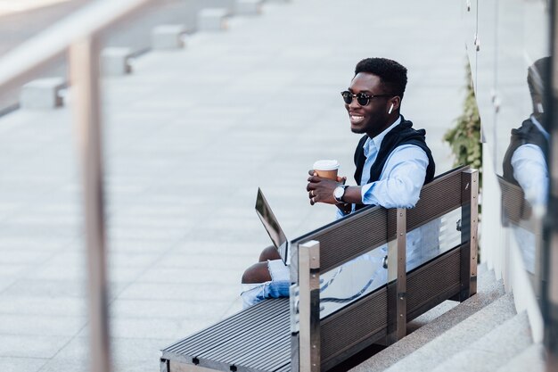 Elegante joven empresario emplazado en un banco con su computadora portátil en una calle soleada junto a un parque. Con taza de café. Estilo de vida.