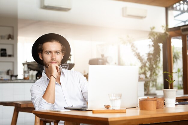 Elegante joven estudiante caucásico con sombrero negro descansando durante el trabajo en el proyecto de diploma, sentado en la mesa de café frente a la computadora portátil abierta, apoyado en su codo y mirando con una sonrisa