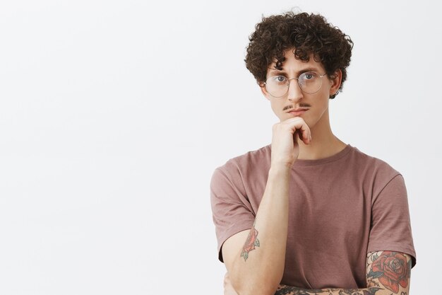 Elegante joven inteligente y creativo con bigote y tatuajes en los brazos con gafas pensando frotarse la barbilla y mirar seriamente