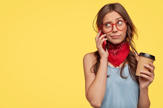 Elegante joven morena con gafas posando contra la pared amarilla