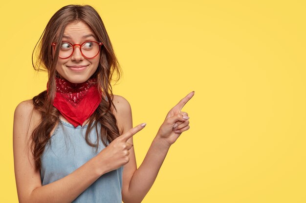 Elegante joven morena con gafas posando contra la pared amarilla