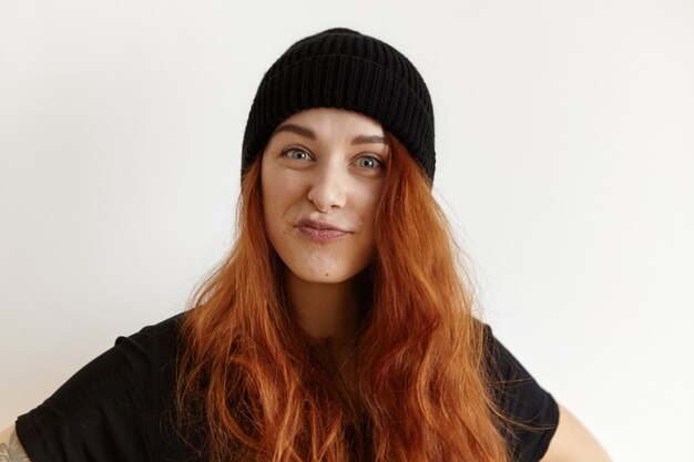 Elegante joven mujer europea con peinado desordenado con sombrero negro y camiseta haciendo pucheros