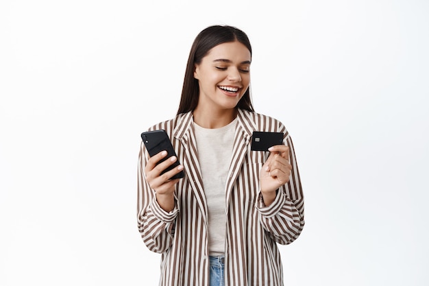Elegante joven mujer moderna sonriendo, mirando a la tarjeta de crédito de plástico, pagando en línea con el teléfono móvil, comprando en la aplicación de teléfono inteligente, pared blanca