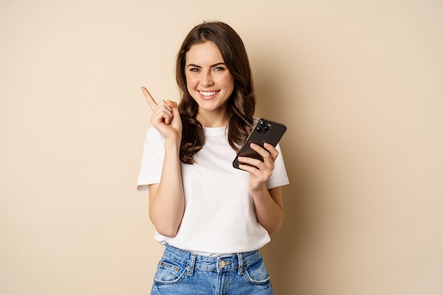 Elegante joven mujer moderna sosteniendo teléfono móvil apuntando con el dedo a la izquierda y sonriendo concepto de compras...