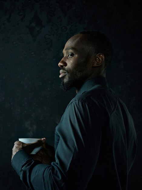 Elegante joven negro africano con taza de café blanca posando sobre fondo oscuro de estudio.