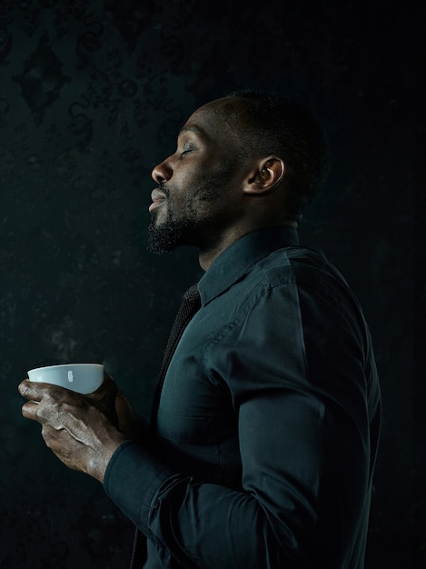 Elegante joven negro africano con taza de café blanca posando sobre fondo oscuro de estudio.