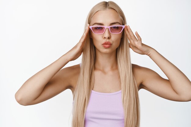 Elegante joven rubia con gafas de sol de moda, labios fruncidos y mirada atrevida, de pie en blanco