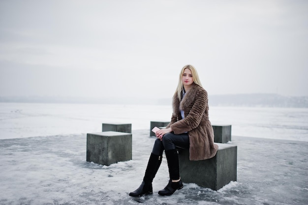 Elegante joven rubia rica en abrigo de piel con teléfono inteligente rosa en cubos de piedra de fondo de mano contra el lago congelado en el día de invierno