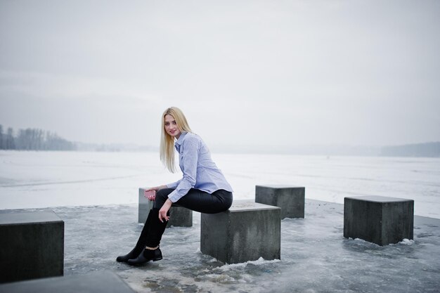 Elegante joven rubia rica en blusa y pantalones de cuero con teléfono inteligente rosa en cubos de piedra de fondo de mano contra el lago congelado en el día de invierno