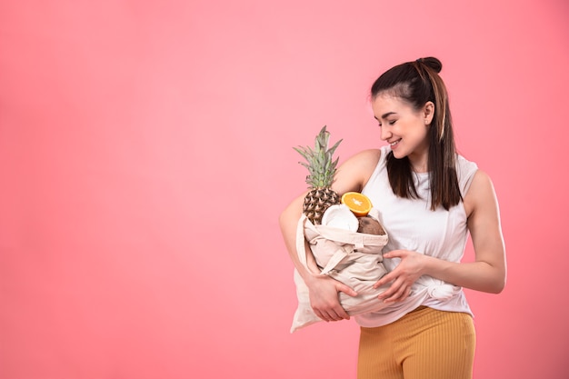 Elegante joven sonriendo y sosteniendo una bolsa ecológica con frutas exóticas en un espacio de copia de fondo rosa.