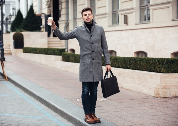 Elegante joven sosteniendo café para llevar en un vaso de papel y coger un taxi en la calle vacía