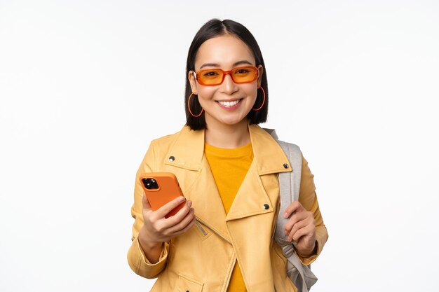 Elegante joven turista asiática con mochila y teléfono inteligente sonriendo a la cámara posando sobre fondo blanco