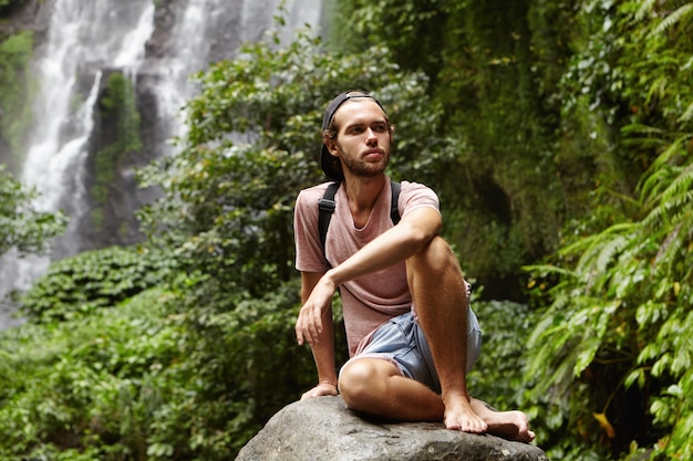 Elegante joven turista caucásico con mochila relajante descalzo en piedra grande con hermosa cascada detrás de él. Mochilero barbudo sentado en la roca durante el viaje en las montañas y con aspecto cansado