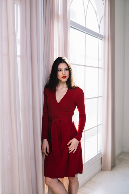 Elegante joven en vestido rojo junto a la ventana en la habitación