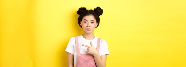 Foto gratuita elegante jovencita asiática con maquillaje y ropa de verano apuntando con el dedo a la derecha y con un aspecto serio.