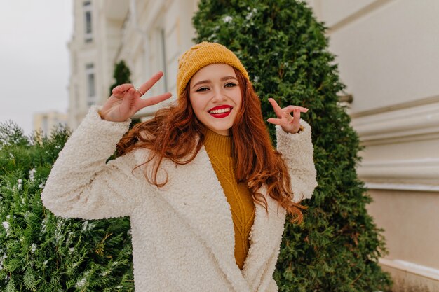 Elegante jovencita jengibre disfrutando del invierno. Chica pelirroja complacida posando con el signo de la paz al aire libre.