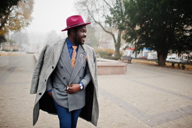 Elegante modelo afroamericano con chaqueta de abrigo gris, corbata y sombrero rojo caminando en la calle con niebla