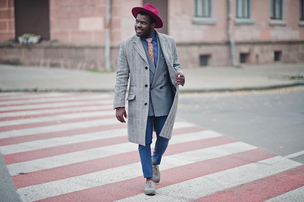 Elegante modelo afroamericano con chaqueta de abrigo gris, corbata y sombrero rojo caminando en el cruce de peatones