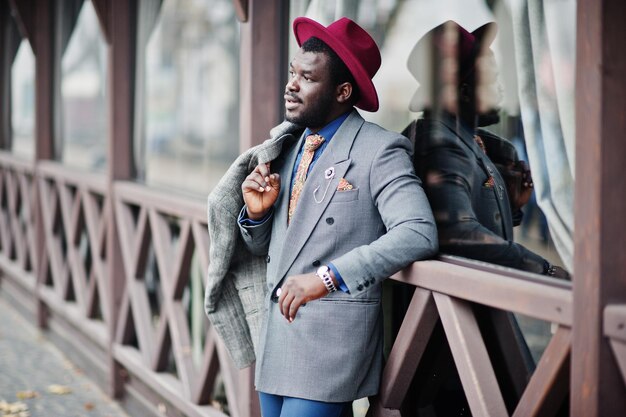 Elegante modelo afroamericano con chaqueta de abrigo gris, corbata y sombrero rojo posado contra un café de madera