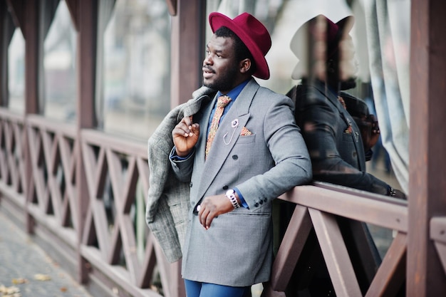 Elegante modelo afroamericano con chaqueta de abrigo gris, corbata y sombrero rojo posado contra un café de madera