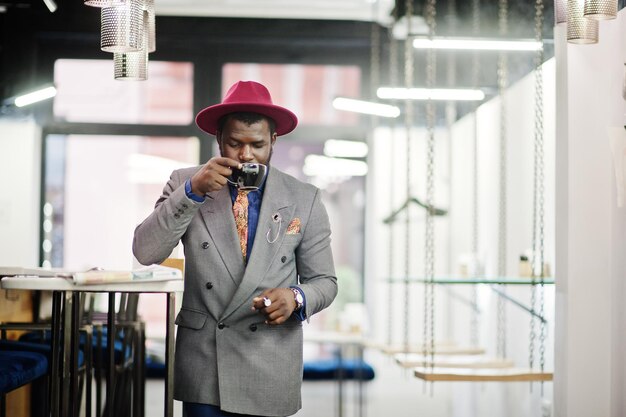 Elegante modelo afroamericano con chaqueta gris, corbata y sombrero rojo, bebe café en el café y lee periódicos