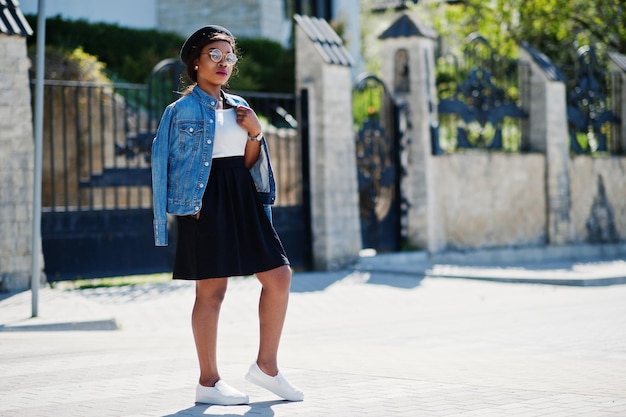 Elegante modelo afroamericano con gafas, sombrero, chaqueta de jeans y falda negra posada al aire libre