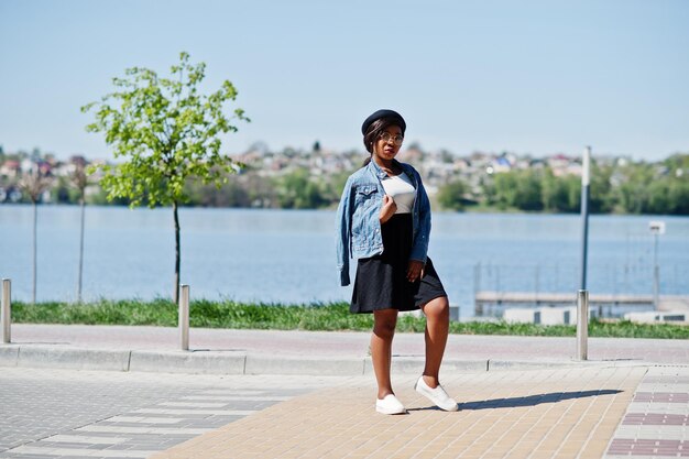 Elegante modelo afroamericano con gafas, sombrero, chaqueta de jeans y falda negra posada al aire libre