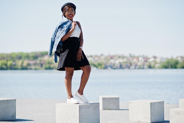 Elegante modelo afroamericano con gafas, sombrero, chaqueta de jeans y falda negra posada al aire libre