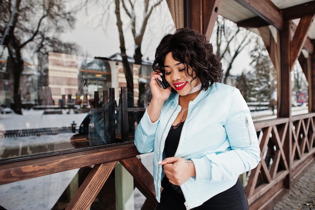 Elegante modelo afroamericano de talla grande con teléfono móvil a mano contra un café de madera en el día de invierno