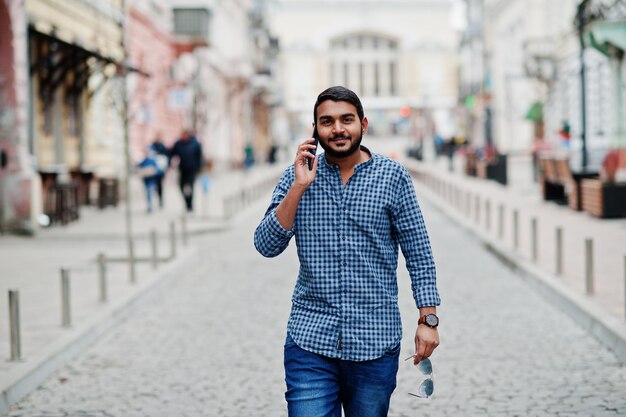 Elegante modelo de barba india con ropa informal posando al aire libre en la calle de la India y hablando por teléfono