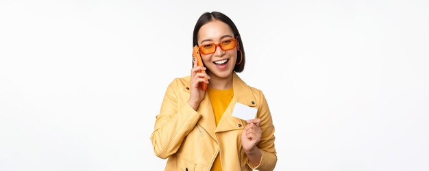 Elegante modelo femenino asiático hablando por teléfono inteligente y mostrando la tarjeta de crédito de pie sobre fondo blanco.