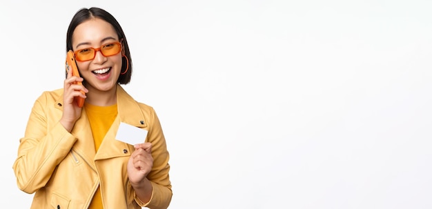 Elegante modelo femenino asiático hablando por teléfono inteligente y mostrando la tarjeta de crédito de pie sobre fondo blanco.