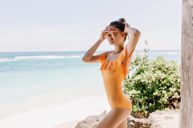 Elegante modelo femenino blanco en ropa amarilla posando en la playa. Chica caucásica delgada en traje de baño naranja escalofriante cerca del mar con los ojos cerrados.