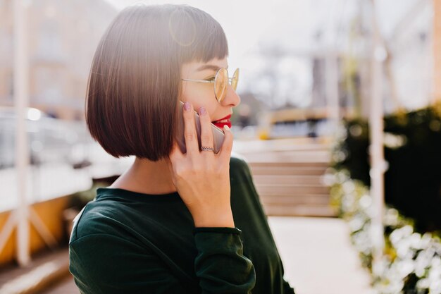 Elegante modelo femenino caucásico en gafas de sol usando su teléfono inteligente en un día soleado Disparo al aire libre de una encantadora mujer morena con suéter verde hablando por teléfono