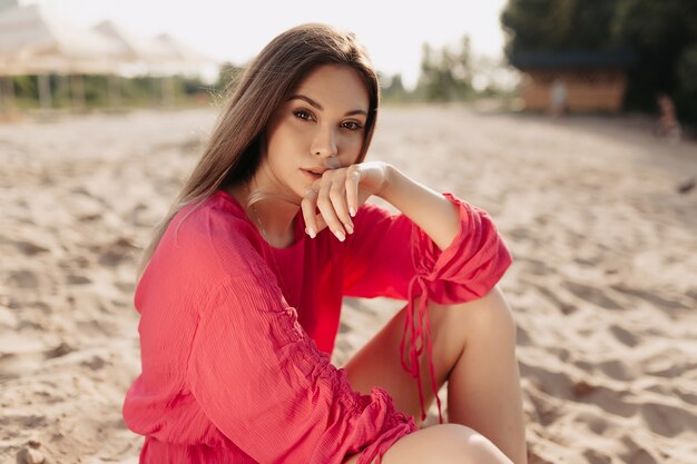 Elegante modelo femenino moderno en vestido rosa de verano posando en la playa en la luz del sol