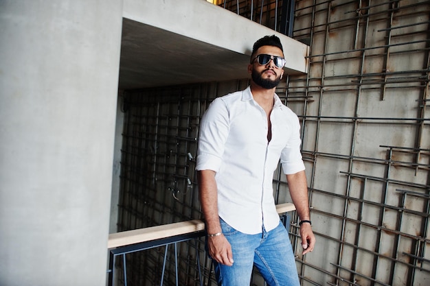 Elegante modelo de hombre árabe alto con camisa blanca, jeans y gafas de sol posados contra la pared de acero interior Barba atractivo chico árabe