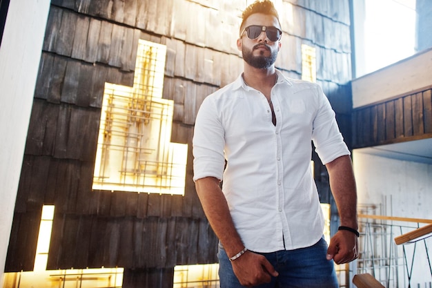 Elegante modelo de hombre árabe alto con camisa blanca, jeans y gafas de sol posados contra la pared de luz interior Barba atractivo chico árabe