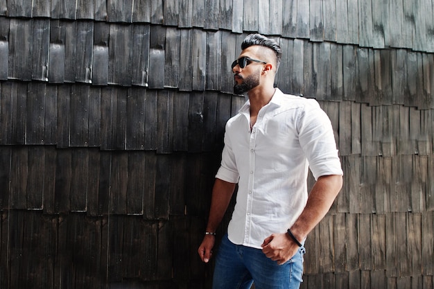 Elegante modelo de hombre árabe alto con camisa blanca, jeans y gafas de sol posados contra la pared de madera interior Barba atractivo chico árabe