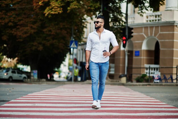 Elegante modelo de hombre árabe alto con pantalones vaqueros de camisa blanca y gafas de sol posados en la calle de la ciudad Barba atractivo chico árabe con una taza de café caminando en un cruce peatonal o en un cruce de peatones