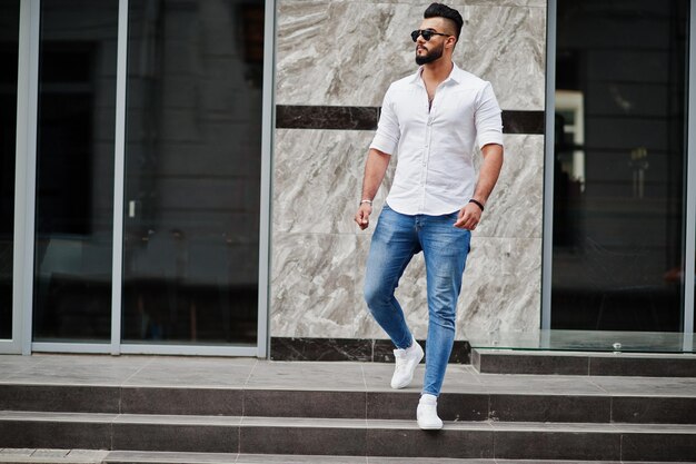 Elegante modelo de hombre árabe alto con pantalones vaqueros de camisa blanca y gafas de sol posados en la calle de la ciudad Chico árabe atractivo con barba