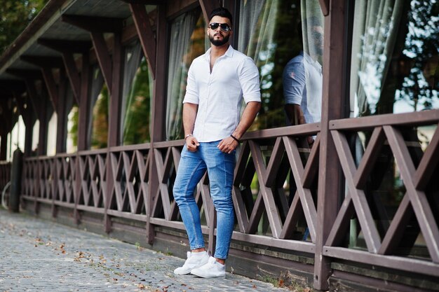 Elegante modelo de hombre árabe alto con pantalones vaqueros de camisa blanca y gafas de sol posados en la calle de la ciudad Chico árabe atractivo con barba