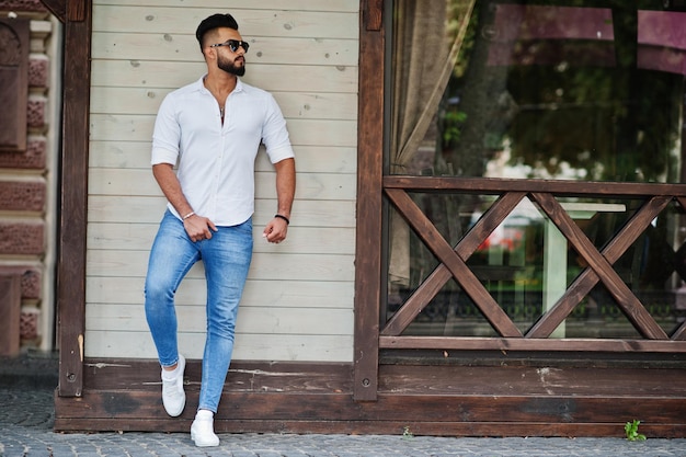 Elegante modelo de hombre árabe alto con pantalones vaqueros de camisa blanca y gafas de sol posados en la calle de la ciudad Chico árabe atractivo con barba