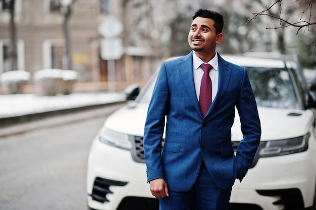 Elegante modelo de hombre de moda indio en traje posado en el día de invierno contra un automóvil suv de negocios blanco
