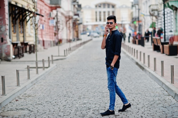 Elegante modelo indio con ropa informal, camisa negra y gafas de sol posadas al aire libre en la calle de la India y hablando por teléfono