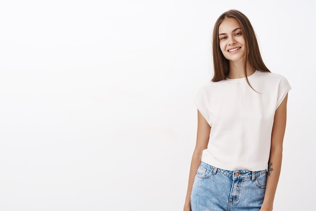 Elegante modelo de mujer adulta segura y atractiva con tatuaje en el brazo posando en una blusa blanca de moda y jeans inclinando la cabeza y sonriendo con mirada despreocupada y amigable