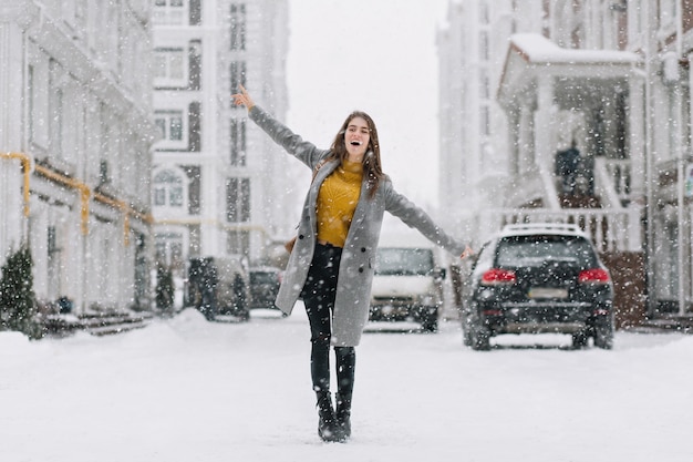 Elegante modelo de mujer caucásica en abrigo largo bailando en la calle en la mañana de invierno. Foto al aire libre de la encantadora dama en suéter amarillo agitando las manos durante la sesión de fotos en un día helado.